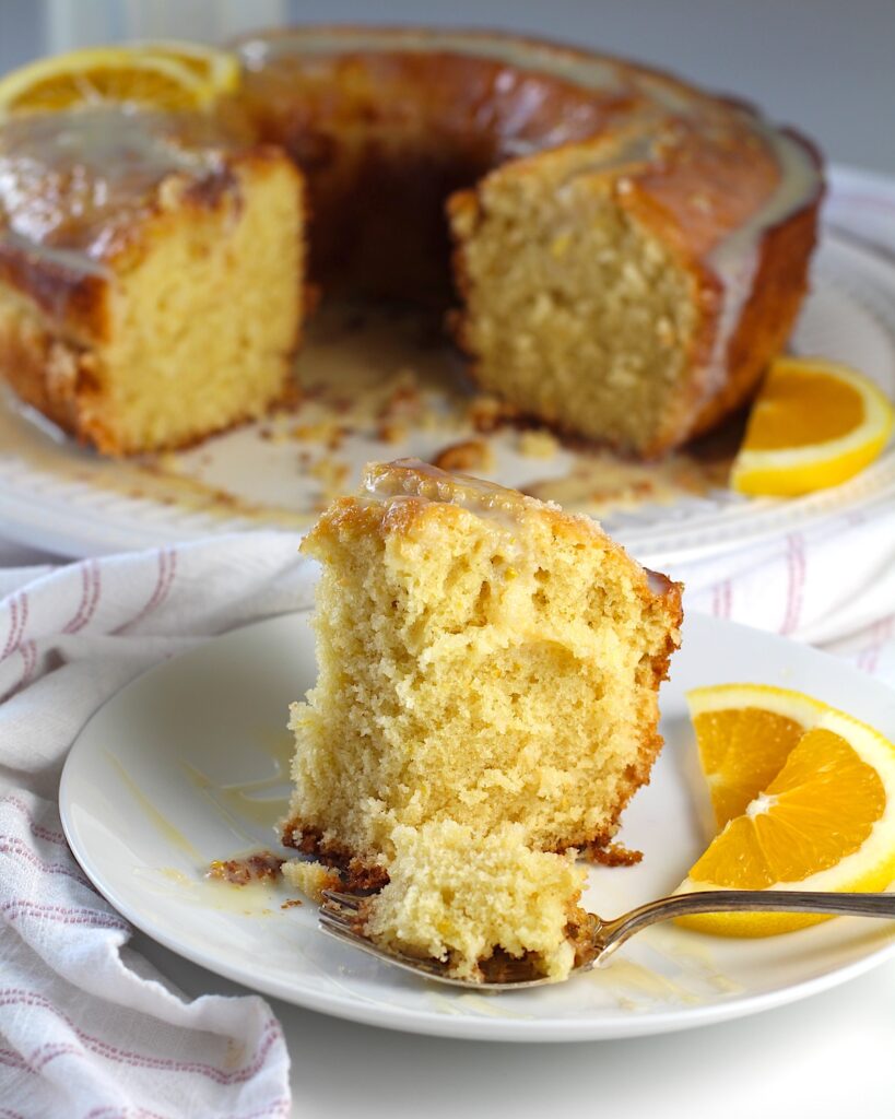 Piece of Orange Bundt Cake (Bolo de Laranja) on a plate with the rest of the cake in background. There are orange slices on the side of the plate and a fork holding a bite on the plate.