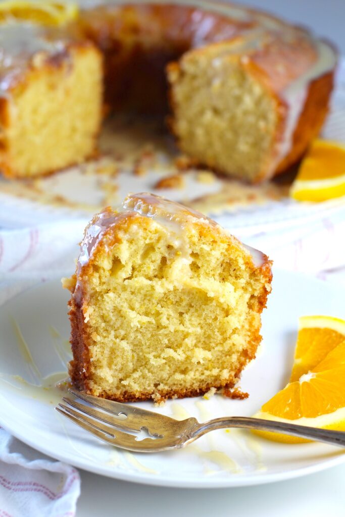 Piece of Orange Bundt Cake (Bolo de Laranja) on a plate with the rest of the cake in background. There are orange slices on the side of the plate and a fork on the plate.