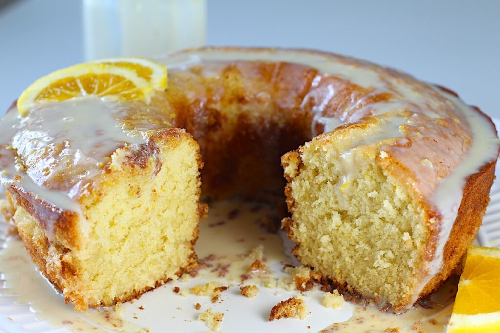 Orange Bundt Cake (Bolo de Laranja) on a platter with front pieces removed, orange slices on top of the cake and on the side of platter and glaze in a clear jar in the background.