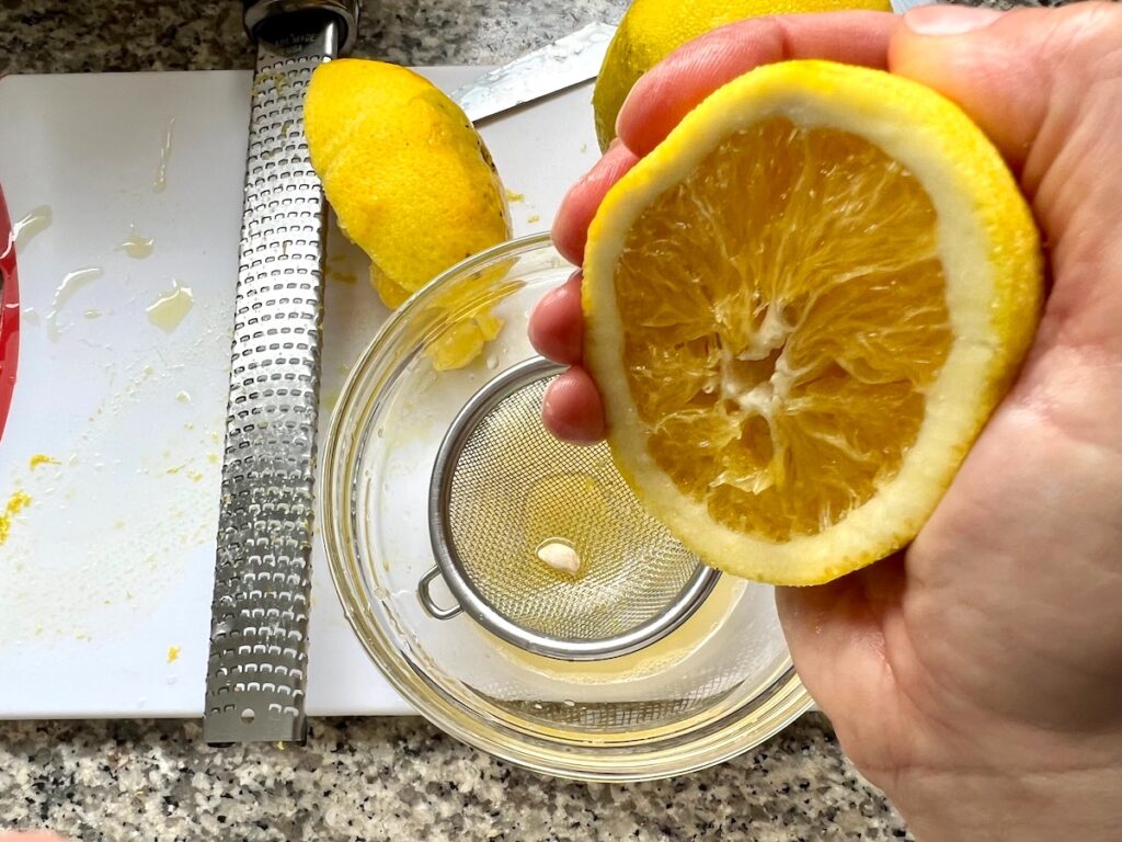 Hand squeezing half of an orange over a small strainer that caught a seed over a bowl on cutting board with microplane next to it for Orange Bundt Cake recipe.