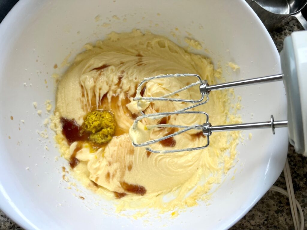 Vanilla extract, orange zest and juice added to batter in a bowl with hand mixer pulled out for Orange Bundt Cake recipe.