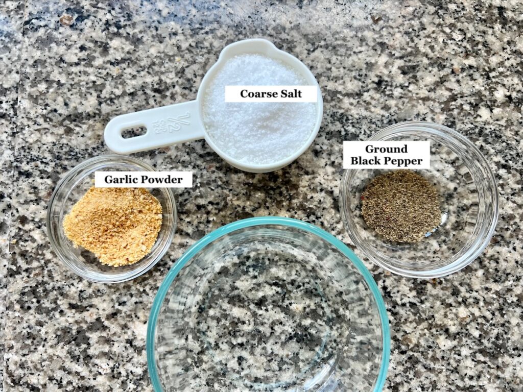 Salt in a measuring cup, garlic powder in a small bowl, black pepper in a small bowl, and a fourth clear empty bowl for the Pork Shoulder Brine.