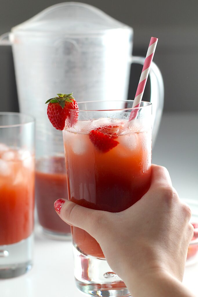 Hand holding a glass of Strawberry Acai drink with pink and white striped straw and strawberries in the glass. In the background is the pitcher of the rest of the drink for this article How to make a strawberry acai refresher.