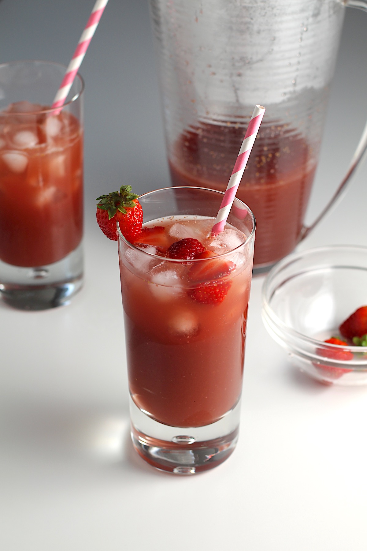Two glasses of Strawberry Acai drink with pink and white striped straws and strawberries in the front glass.  In the background is the pitcher of the rest of the drink for this article How to make a strawberry acai refresher.