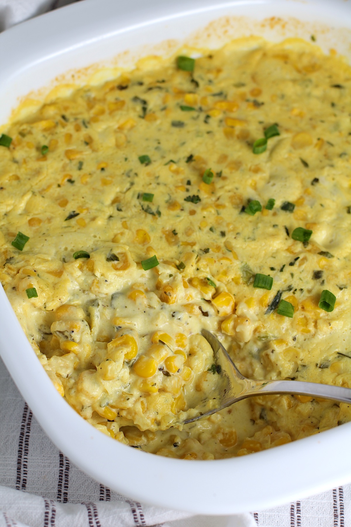 Corn Casserole Recipe with Cream Cheese and scallion slices on top in casserole dish with a spoon scooping some from the corner.