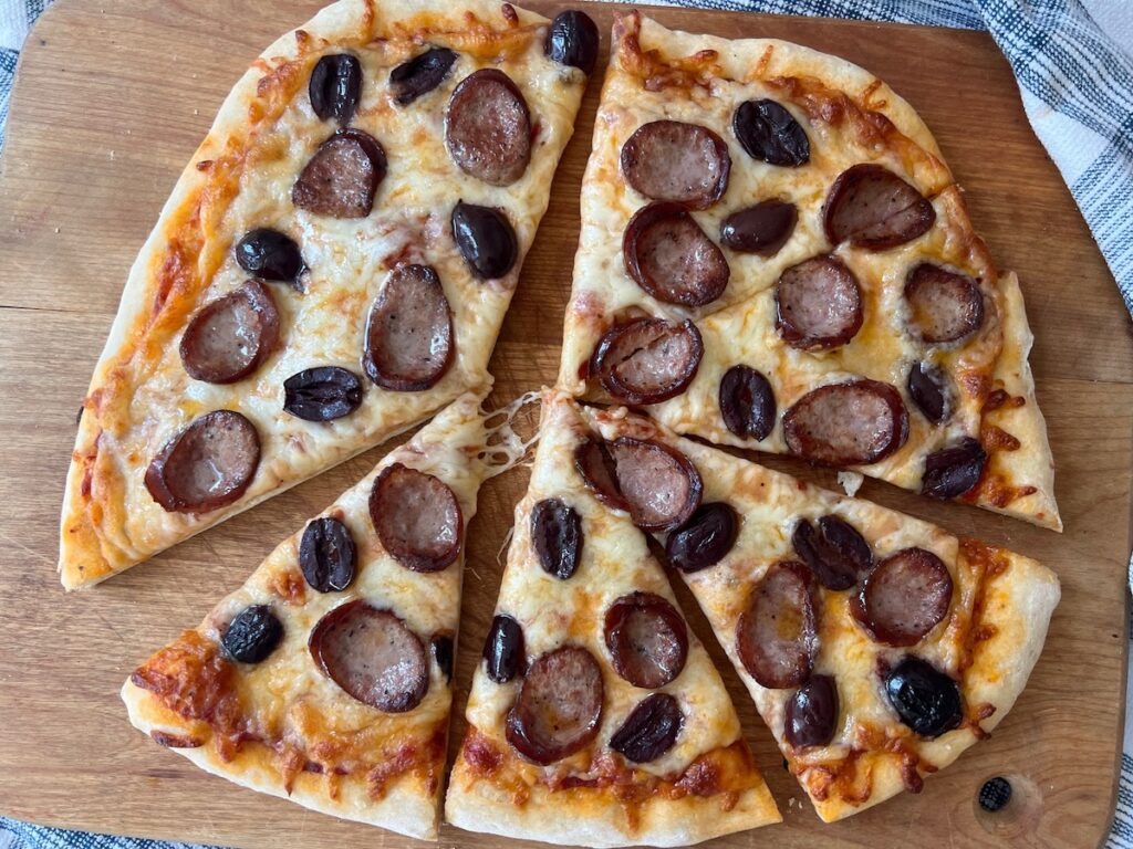 Brazilian Pizza cut into triangles on a wood cutting board on blue and white towel on table. It's a thin, crispy crust topped with mozzarella cheese, cabresa sausage, and olives.