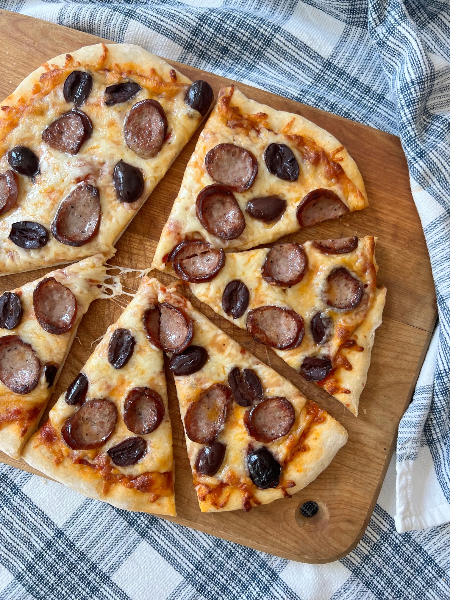 Brazilian Pizza cut into triangles on a wood cutting board on blue and white towel on table.  It's a thin, crispy crust topped with mozzarella cheese, cabresa sausage, and olives.