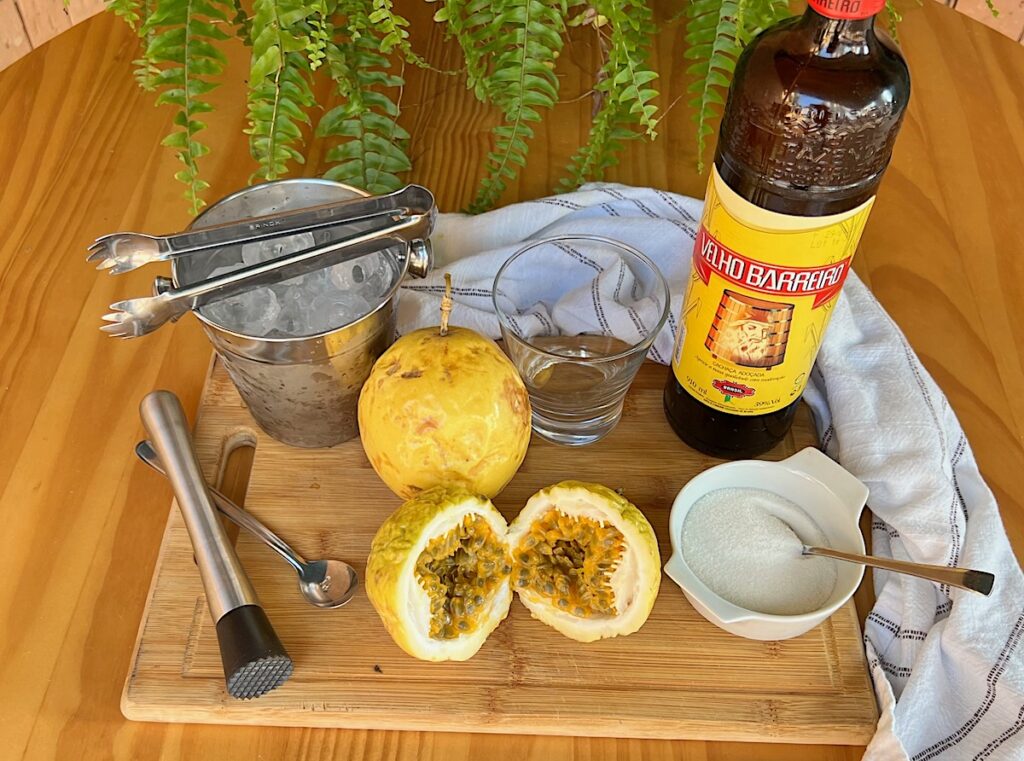 Yellow passion fruit cut in half with inside pulp showing on a cutting board with another whole fruit behind it, next to a martini glass, bottle of cachaca, bowl of sugar, muddler, spoon, bucket of ice on a cutting board for a Passion Fruit Caipirinha recipe.
