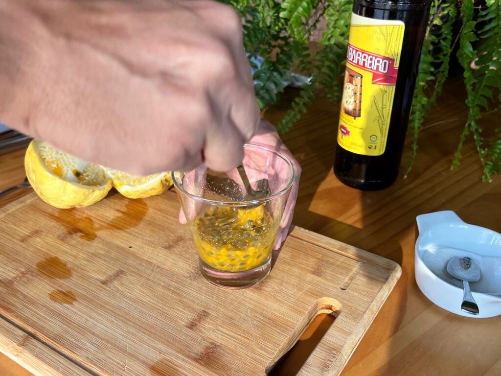 Hand mixing sugar into passion fruit pulp with seeds in a martini glass on a cutting board with a bottle of cachaca in background for a Passion Fruit Caipirinha recipe.