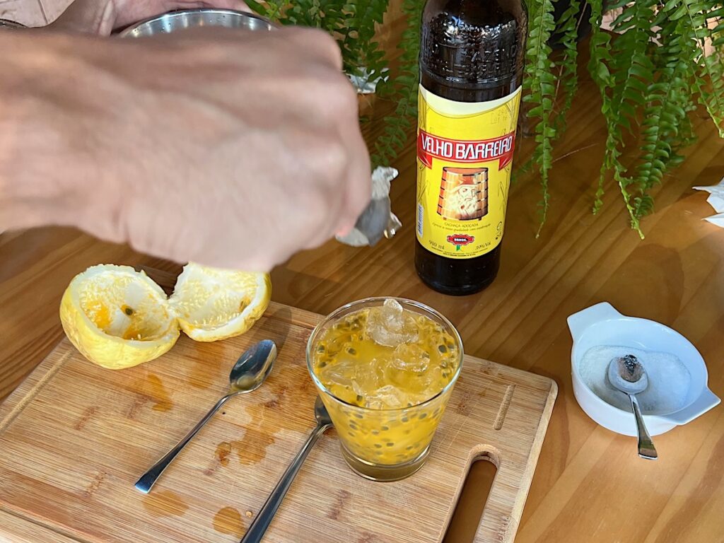 Hand adding ice cubes to a glass of Passion Fruit Caipirinha in a martini glass on a cutting board with cut yellow passion fruit to the left and a bottle of cachaca in background.