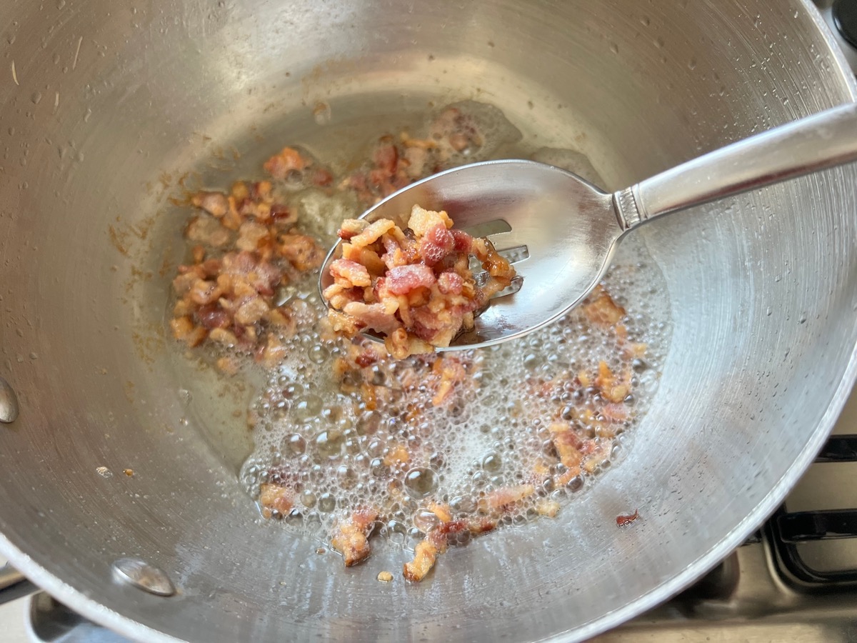 Slotted spoon scooping crisp bacon bits out of bacon fat in a pot for Arroz Pilaf.