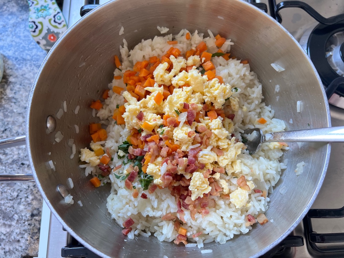 Cooked diced carrots, crisp bacon bits, chopped cilantro, and scrambled eggs added to cooked rice in pot for Arroz Pilaf.