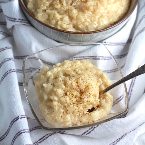 Rice Pudding with condensed milk in a clear serving bowl with cinnamon on top and a spoon scooping. A larger bowl of rice pudding is in the background.