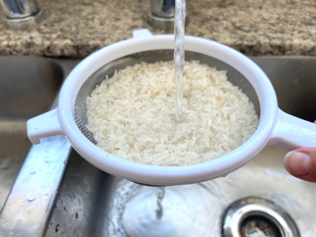 Uncooked white rice being rinsed in a strainer under faucet water for Rice Pudding with condensed milk.
