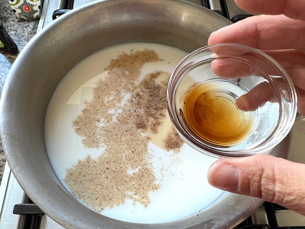 Hand about to pour vanilla extract into milks and white rice in a pot for Rice Pudding with condensed milk.