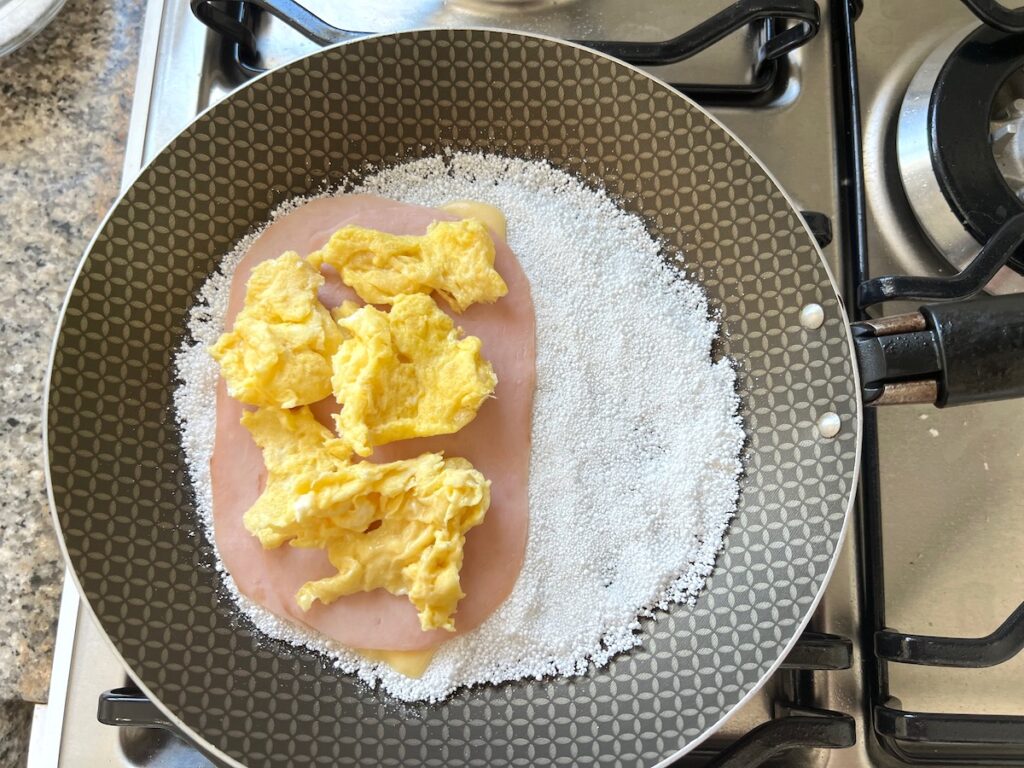 Scrambled eggs on top of ham slices layered onto half of the tapioca starch crepe in a frying pan for Brazilian Breakfast Tapioca recipe.