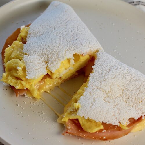 Brazilian Breakfast Tapioca crepe filled with ham, egg, and cheese cut in half with melted cheese pulling in the center on the plate.