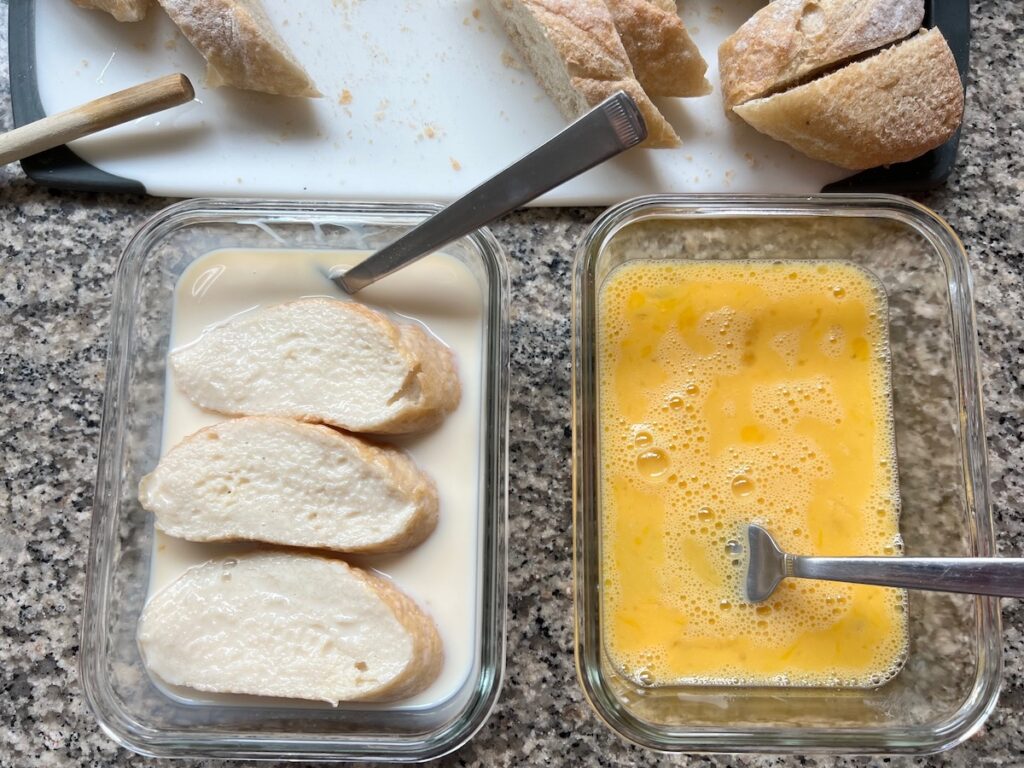 Three slices of baguette soaking in the milk mixture in a dish next to a second dish with beaten eggs and a cutting board in background with baguette slices for Brazilian French Toast with Condensed Milk.