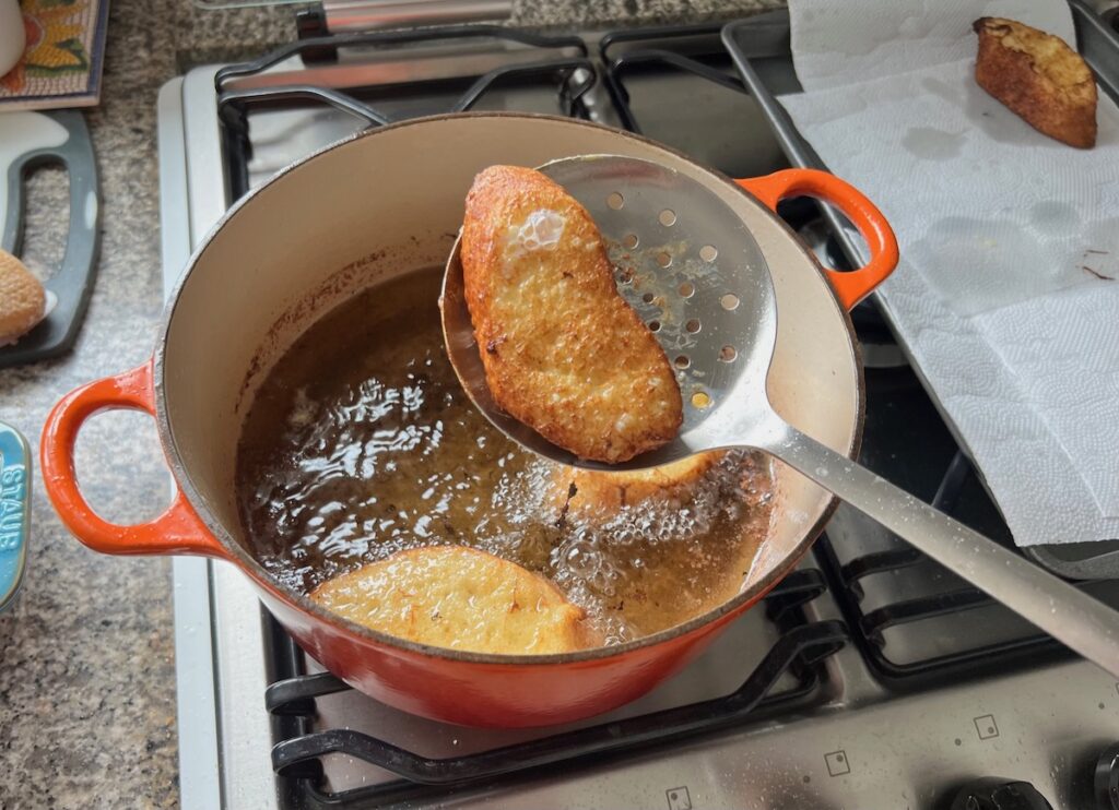 Slotted large flat spoon holding an fried Rabanada, slice of bread over hot oil in pot for Brazilian French Toast with Condensed Milk.
