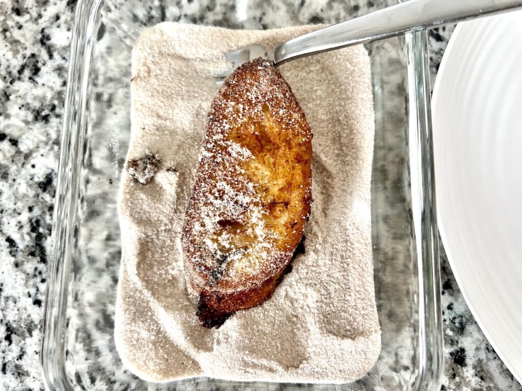 Hot fried Rabanada, slice of french toast, in a shallow dish with cinnamon sugar as a spoon coats all sides for Brazilian French Toast with Condensed Milk.