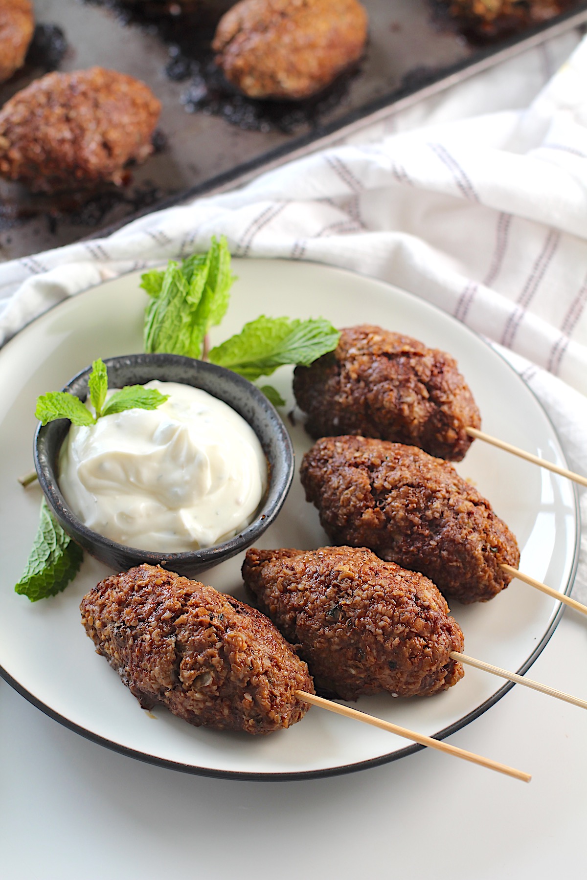Four Kibe kabobs, Mediterranean Ground Beef Kabob recipe, cooked, skewered, and plated with yogurt dipping sauce and fresh mint leaves as garnish.