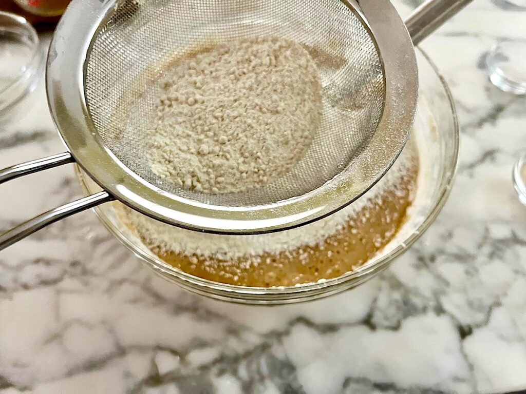 Flour being sifted into batter in a clear glass bowl for the Honey Loaf recipe.