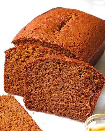 Honey Loaf cake baked beautifully golden brown on table with 3 slices standing in front showing the moist inside. On the counter is a small bowl of honey.