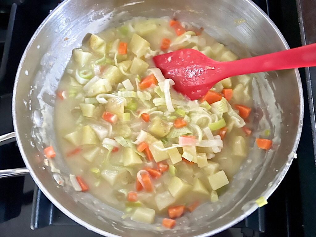 Red spatula stirring broth into to carrots, potatoes, leeks and flour in a pot for the filling for the Ham and Leek Pie.