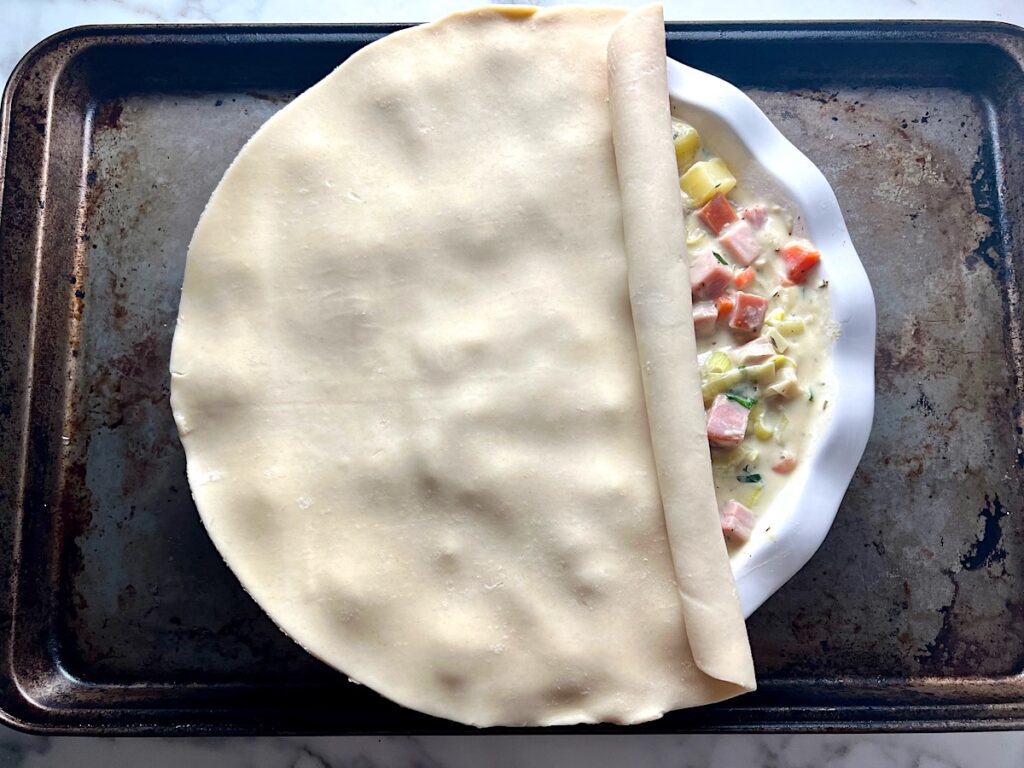 Premade Pie crust dough being rolled out over the filling in a pie dish for Ham and Leek Pie recipe.
