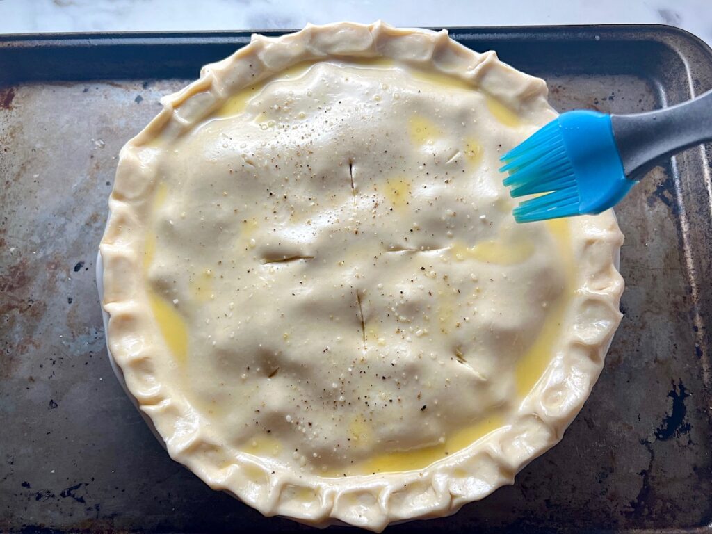 Silicone brush brushing egg wash onto raw pie crust dough with crimped edges and slits on top covering the filling in a pie dish for Ham and Leek Pie recipe.