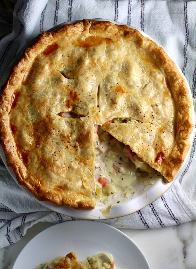 Ham and Leek Pie with potatoes and carrots in a cream sauce and flaky golden brown pie crust on top. Pie in pie pan on counter with 1 slice removed showing creamy, savory filling.