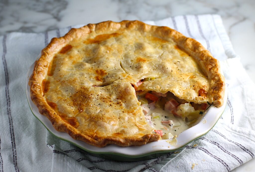 Ham and Leek Pie with potatoes and carrots in a cream sauce and flaky golden brown pie crust on top. Pie in pie pan on counter with 1 slice removed showing creamy, savory filling.