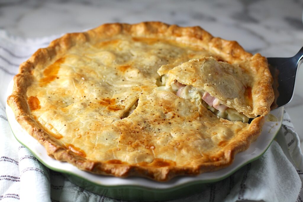 Spatula scooping out a slice of Ham and Leek Pie with potatoes and carrots from the pie dish on a counter.