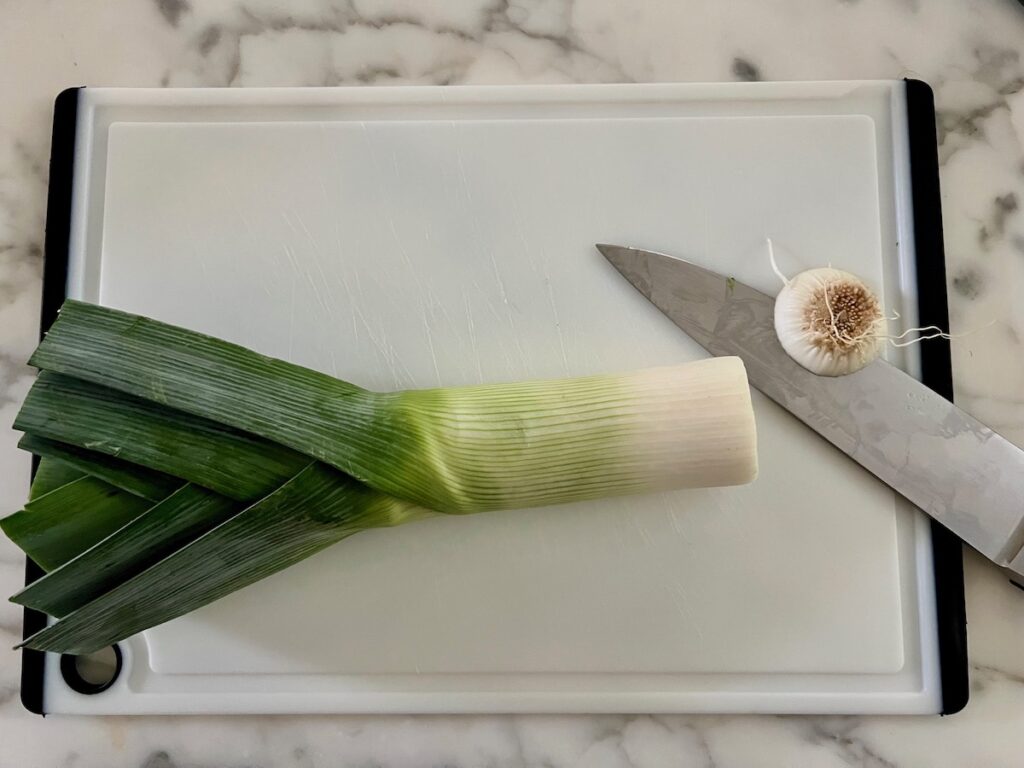 Raw whole leek on cutting board with chefs knife that cut off the root end for the Ham and Leek Pie.