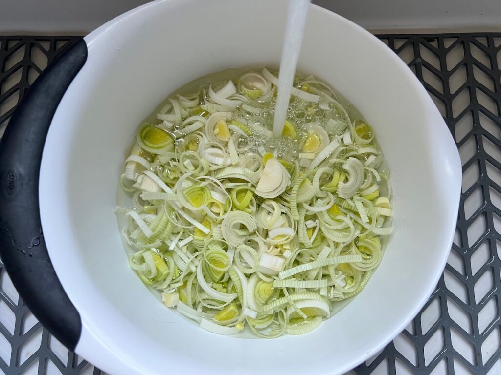 Raw leek slices in a bowl with water with running water pouring into the bowl for the Ham and Leek Pie.