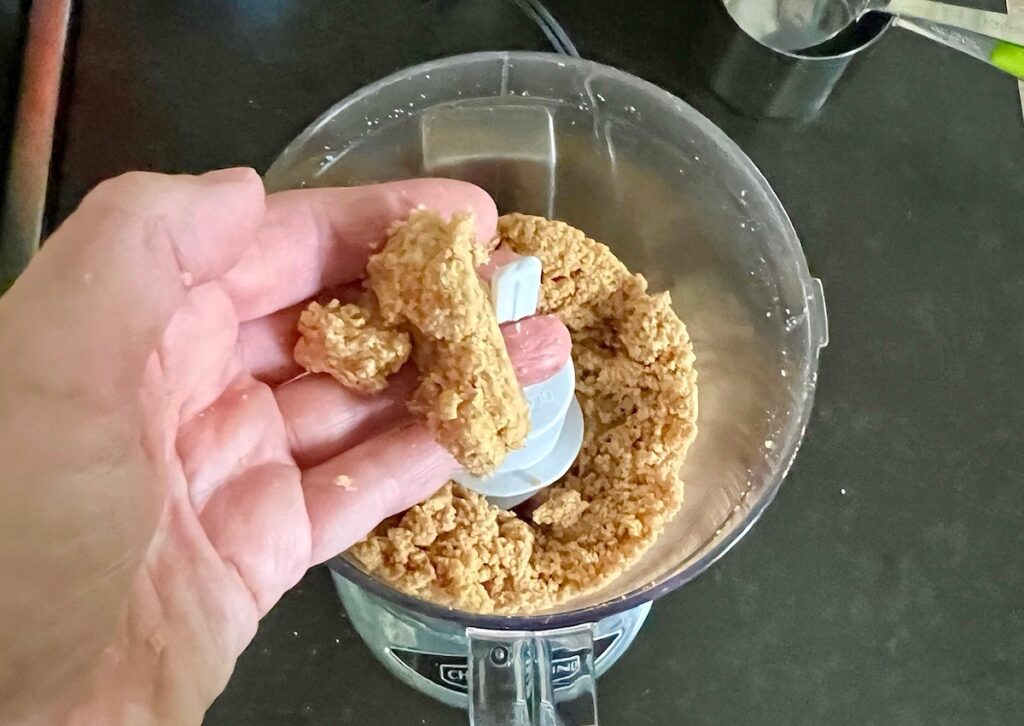 Hand holding some blended peanuts and sugar to show pasty texture over the food processor being blending for Brazilian Peanut Candy recipe (Pacoca).