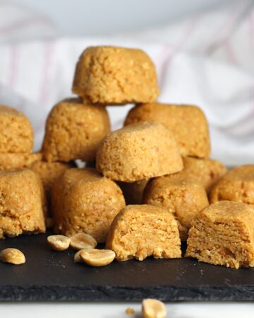 Twelve small mounds of Brazilian Peanut Candy or This Paçoca de Amendoim, stacked on a black platter with one cut open in the front, peanut halves on the platter and table, and a towel in the background.