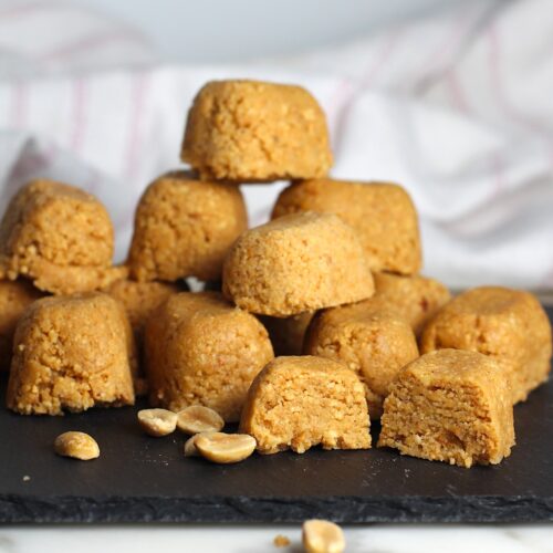 Twelve small mounds of Brazilian Peanut Candy or This Paçoca de Amendoim, stacked on a black platter with one cut open in the front, peanut halves on the platter and table, and a towel in the background.
