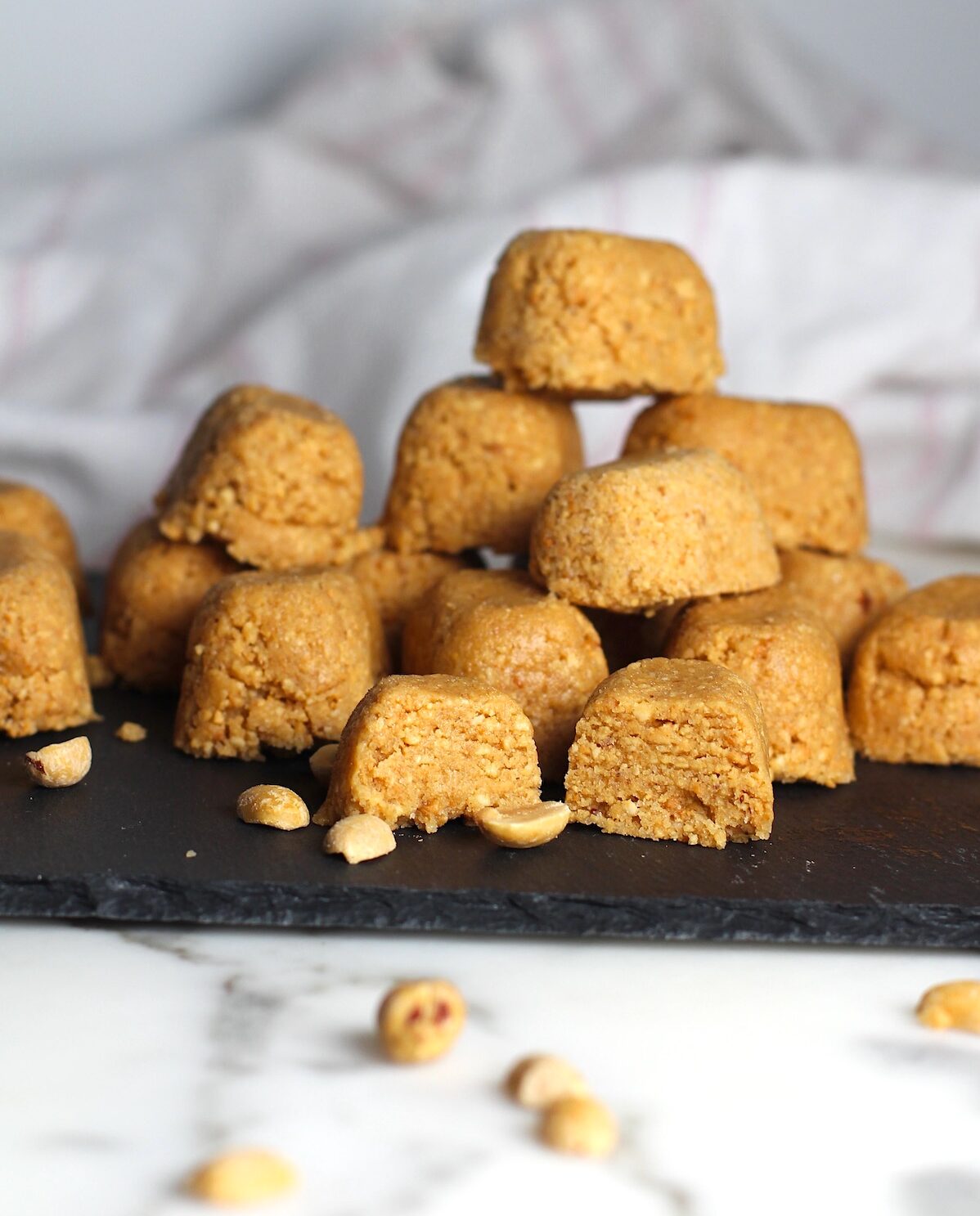 Twelve small mounds of Brazilian Peanut Candy or This Paçoca de Amendoim, stacked on a black platter with one cut open in the front, peanut halves on the platter and table, and a towel in the background.