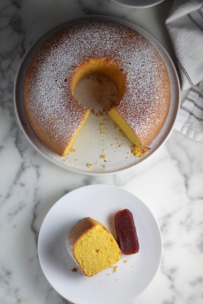 Piece of Bolo de Fuba (cornmeal Bundt Cake) on white plate with slice of guava paste and whole cake with missing piece in background.