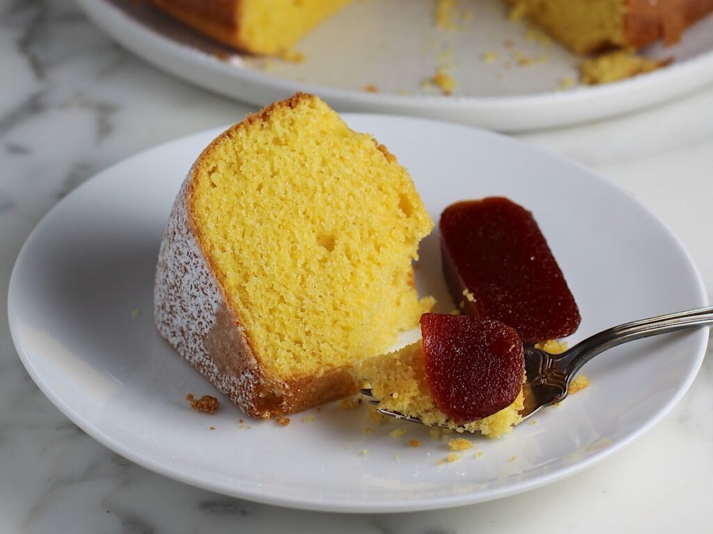 Piece of Bolo de Fuba (cornmeal Bundt Cake) on plate with fork holding a bite with guava paste on top and on the side.  The whole cake with missing piece is in background.