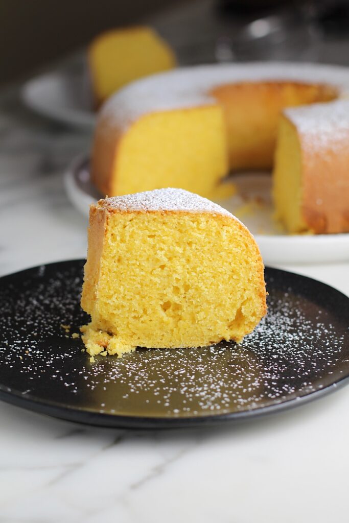 Piece of Bolo de Fuba (cornmeal Bundt Cake) on plate with powdered sugar dusted on top and whole cake with missing piece in background.