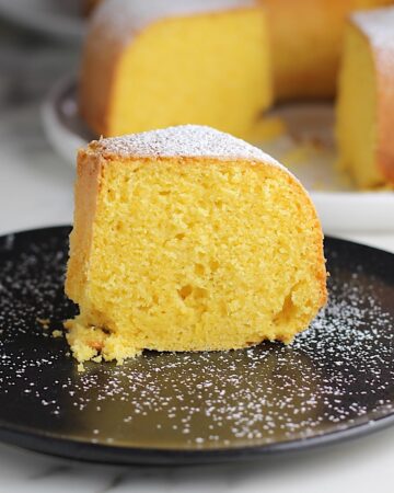 Piece of Bolo de Fuba (cornmeal Bundt Cake) on black plate with powdered sugar dusted on top and whole cake with missing piece in background.