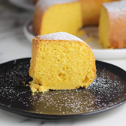 Piece of Bolo de Fuba (cornmeal Bundt Cake) on black plate with powdered sugar dusted on top and whole cake with missing piece in background.
