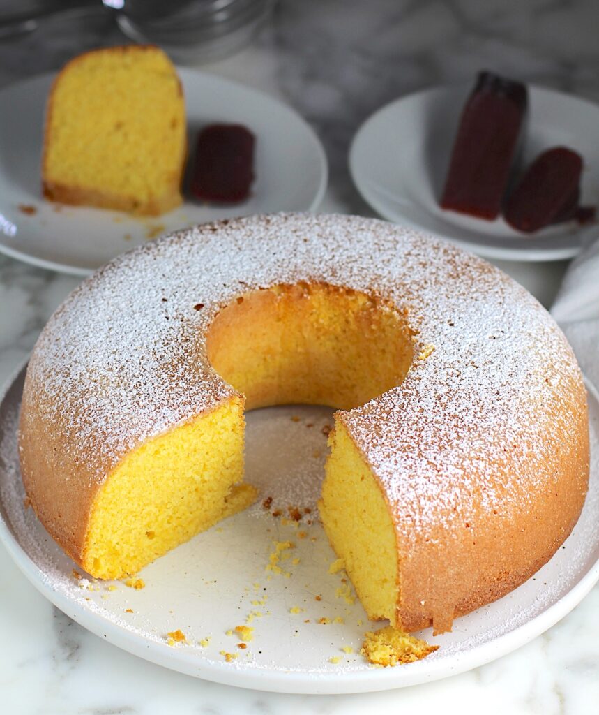 Bolo de Fuba (cornmeal Bundt Cake) on plate with powdered sugar dusted on top and large piece removed from the front of the bundt cake.  Slice of cake on a plate in the back and guava paste on another plate in the back.