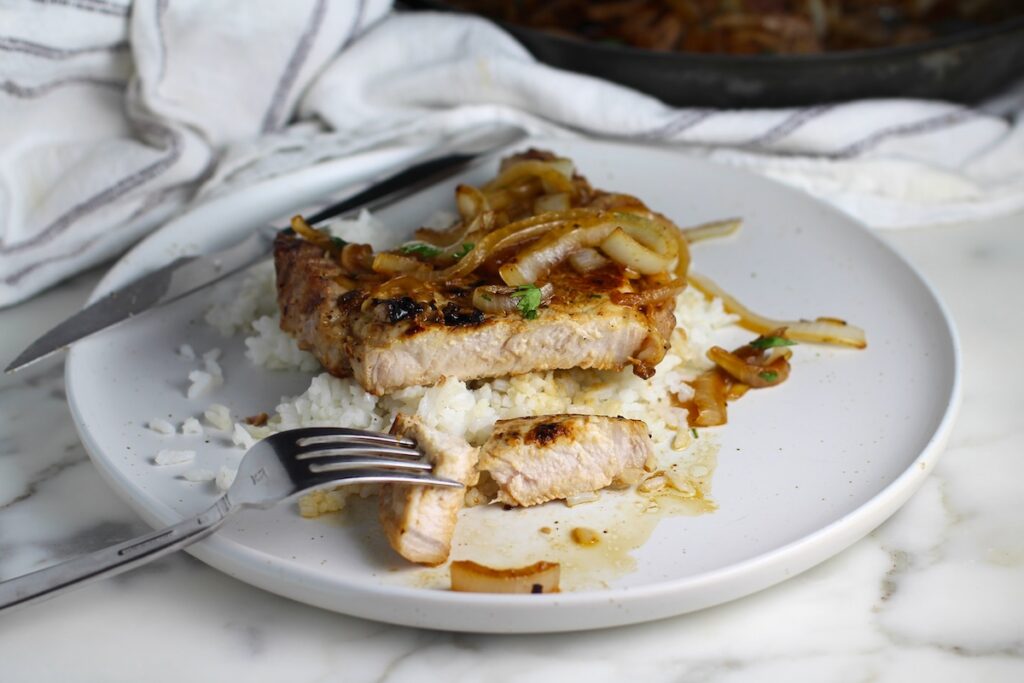Browned cooked bone-in pork chop Sliced with fork holding a bite.  The pork is topped with sauteed onions all over white rice on a plate for this Pork Chops with Onions recipe, Costeletas de Porco com Cebolas.