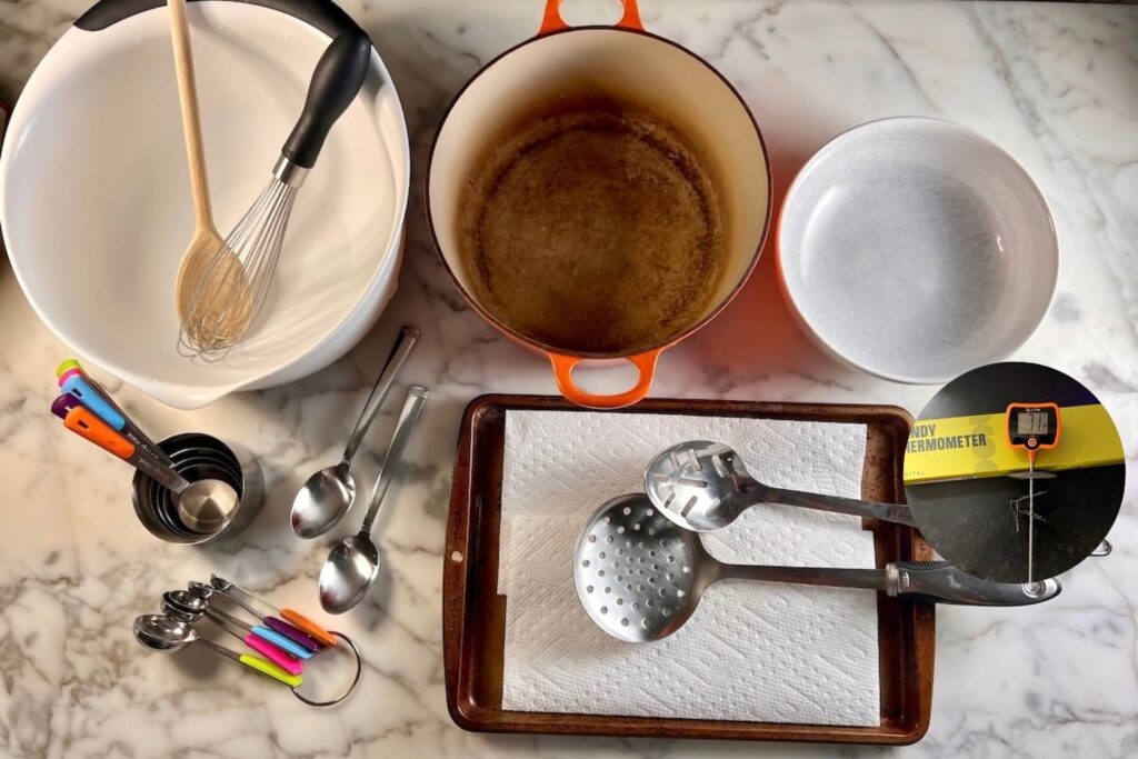 Equipment out on counter for the Bolinhos de Chuva Recipe (Donut holes with Cinnamon Sugar coating). Includes a large mixing bowl with whisk and wood spoon, a Dutch oven, candy thermometer, wide shallow bowl for cinnamon sugar, slotted spoon, 2 soup spoons, measuring spoons, and measuring cups.