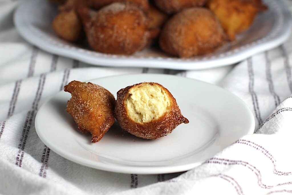On a small plate, a bite is taken out of a Bolinho de Chuva (Donut hole with Cinnamon Sugar coating) showing the moist inside.  In the background is the plate with more bolinhos stacked on it.  Plates are on the marble counter with blue and white striped towel under.