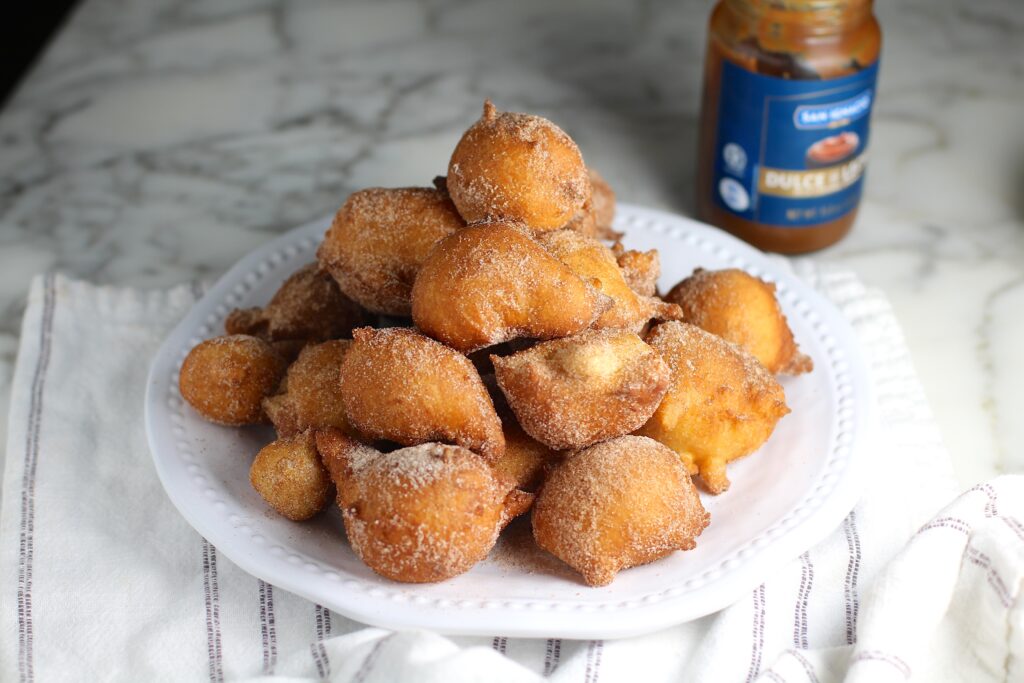 Side view of Bolinhos de Chuva (Donut holes with Cinnamon Sugar coating) stacked on a plate in a pyramid with 1 donut on top. Plate is on the marble counter with Dulce de leche jar in background and blue and white striped towel under.