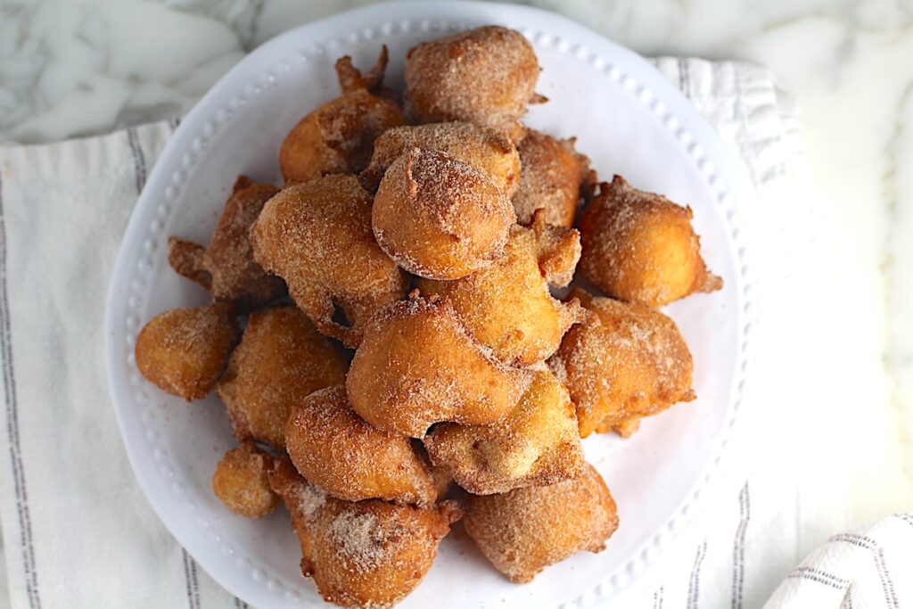 Overhead view of Bolinhos de Chuva (Donut holes with Cinnamon Sugar coating) stacked on a plate in a pyramid with 1 donut on top. Plate is on the marble counter with blue and white striped towel under.